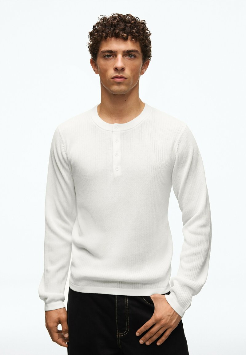 Jeune homme aux cheveux bouclés portant une chemise henley blanche à manches longues et un pantalon noir, debout avec une main dans la poche.