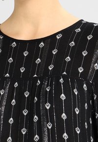 Schwarzes Stoffoberteil mit silberner Blumenstickerei in vertikalen Linien, einem runden Ausschnitt und weicher Textur. Elemente umfassen geraffte Details.