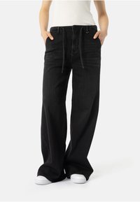 Cilvēks valkā plašas melnas džinsus ar aukliņu jost�ā un baltas kedas, rokas kabatās, pret gludu fona.