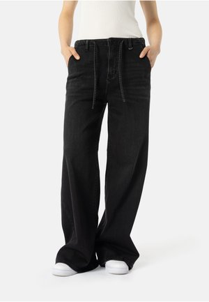 Person med vide sorte jeans med snor i taljen og hvide sneakers, hænderne i lommerne, foran en ensfarvet baggrund.