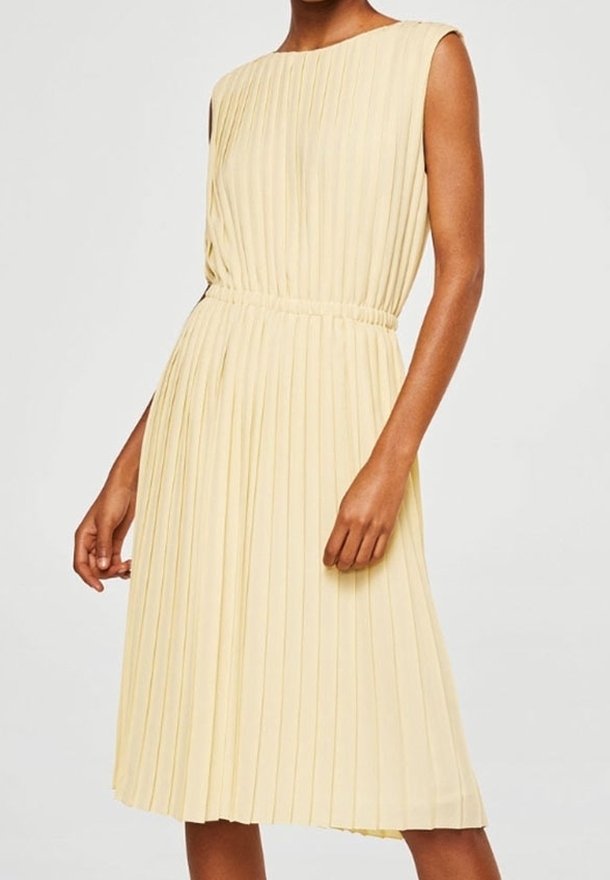 Robe crème plissée sans manches, longueur genou, avec taille cintrée, portée par une personne debout devant un fond uni.
