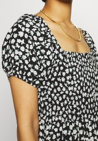 Vestido negro floral con patrón blanco y verde, mangas abullonadas, escote cuadrado y cuerpo fruncido. Collar de cadena dorada como acento.