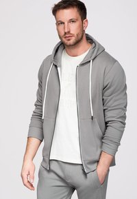 Homme portant un sweat à capuche gris zippé sur un t-shirt blanc et un pantalon gris assorti, debout avec une main dans la poche sur un fond uni.