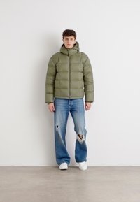 Polstret olivengrønn jakke med hette, med glidelås og quiltet design, brukt med slitte blå jeans og hvite sneakers.