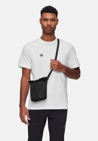 Svart axelremsväska med dragsko, tillverkad av slitstarkt tyg; bärs över en vit t-shirt med ett runt tryck och rundad hals.