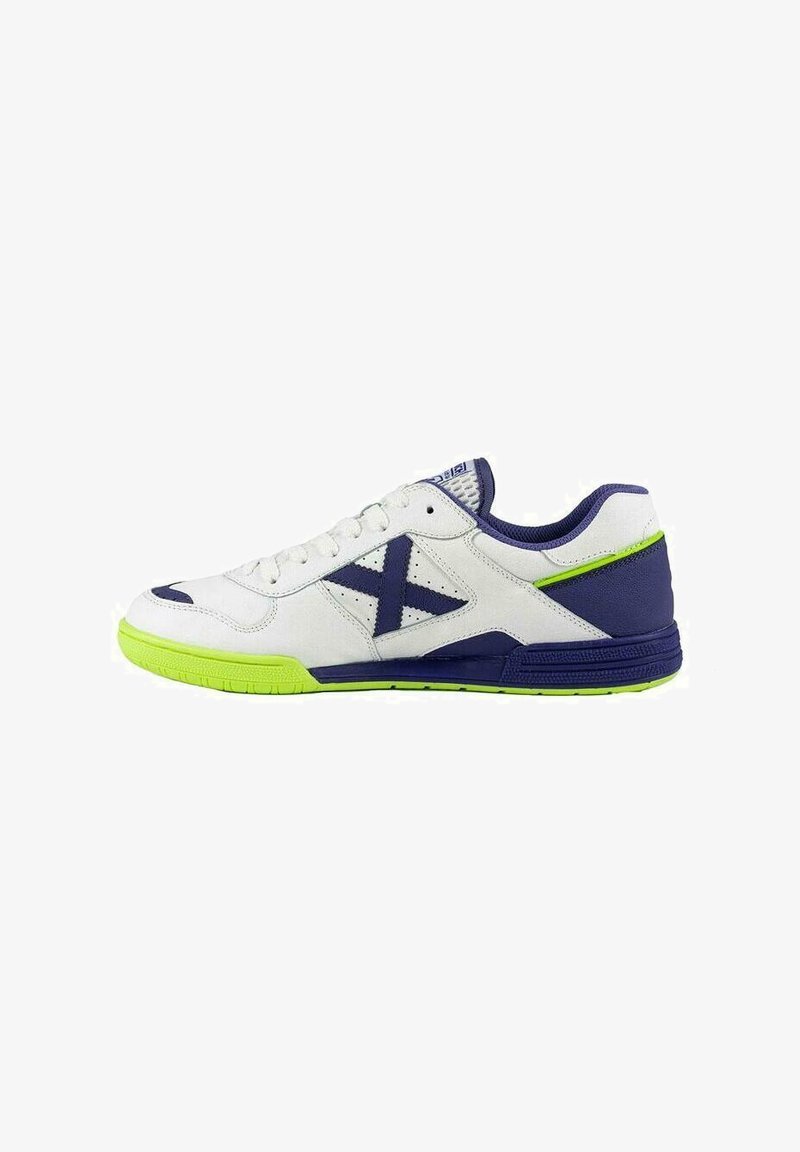 Zapatilla deportiva blanca con acentos en azul marino y suela de color verde brillante. Presenta un upper liso, punta redondeada y parte frontal con cordones, además de detalles perforados.
