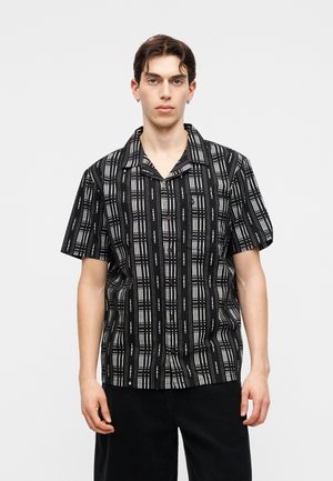 Νέος άνδρας με σκούρα μαλλιά, ντυμένος με κοντομάνικο πουκάμισο με μαύρο και άσπρο μοτίβο και μαύρο παντελόνι, στέκεται μπροστά σε απλό φωτεινό φόντο.