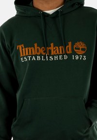 Sudadera verde hecha de tela suave, con el logo de "Timberland" en naranja y blanco, un bolsillo tipo canguro y capucha ajustable con cordón.