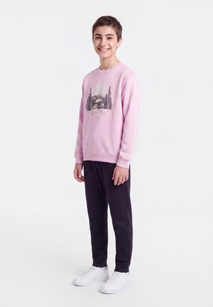 Adolescent garçon debout et souriant, portant un sweat rose avec un motif de montagne, un pantalon noir et des baskets blanches sur fond blanc.