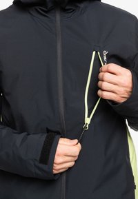 Schwarze Jacke mit Kapuze, ausgestattet mit einem gelben Reißverschluss auf der Brust und einem Klettverschluss am Ärmel. Texturiertem Stoff und aerodynamischem Design für Wärme.