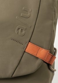 Borsa verde oliva con tessuto lavorato, dotata di una tracolla in pelle marrone e logo impresso. Dettaglio di cuciture visibile lungo il bordo.