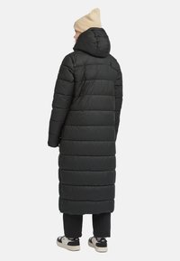 Zwarte pufferjas met een lange, oversized ontwerp en horizontale stiksels, voorzien van een capuchon en een gladde textuur. Gecombineerd met zwarte broek en sneakers.