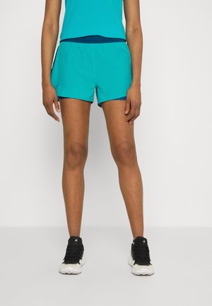 Personne portant un short de sport bleu sarcelle, un haut assorti et des chaussures de course noires et blanches, debout sur un fond blanc.