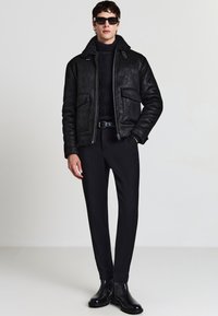 Uomo che indossa un giubbotto in shearling nero, dolcevita a maglia nera, pantaloni neri su misura, cintura nera, stivali neri e occhiali da sole su uno sfondo neutro.