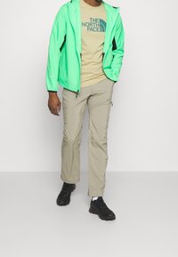 Chaqueta ligera verde neón con detalles en negro, camiseta beige con un logo, pantalones cargo color canela y zapatillas negras.