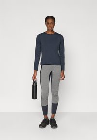 Námornícky atletický top s dlhým rukávom skombinovaný so sivými legínami s námorníckymi akcentmi. Model drží čiernu fľašu na vodu a má na sebe čierne tenisky.