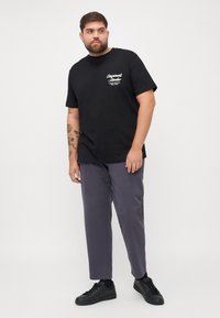Schwarzes Baumwoll-T-Shirt mit aufgedrucktem Logo auf der Brust, kombiniert mit lila Hosen und schwarzen Sneakers. Das Model posiert vor einem neutralen Hintergrund.