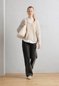 Beige strikkegenser over en hvit skjorte med krage, mørke vide bukser og lyse sko. En kremfarget veske holdes i høyre hånd.