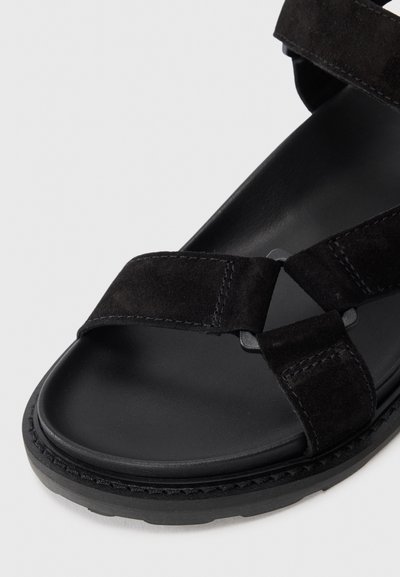 Sandale negre din suede cu curele late, talpă căptușită și talpă exterioară din cauciuc texturat. Caracterizează un design cu curele încrucișate și accente hardware minime.