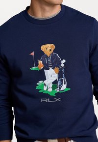 Marinblå sweatshirt med en bild av en björn i golfkläder som håller en drink, med en golfbag och en flagga i bakgrunden. "RLX"-logotyp på framsidan.