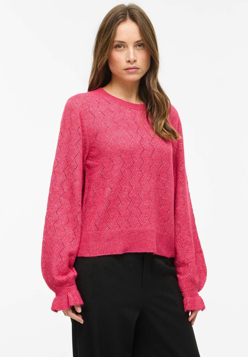 Femme portant un pull en tricot rose avec des manches bouffantes et un pantalon noir, debout devant un fond blanc uni.