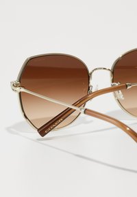 Brune gradientlinser solbriller med gullmetallramme, firkantet form, og brune acetatstenger med "COACH"-logo.