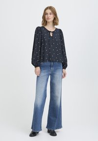 Blusa de manga larga azul oscuro con estampado de corazones blancos, escote en forma de llave y mangas abullonadas. Combinada con jeans de pierna ancha azules y botas negras.