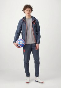 Blauer Sportjacke mit orangefarbenen Akzenten, grauem Shirt und passender Hose. Hält einen mehrfarbigen Fußball, trägt weiße Stollenschuhe.