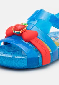 Blå gjennomsiktige sandaler med en rød sløyfe og epledekorasjon; tåle og teksturert såle har et fargerikt Snøhvit-mønster.