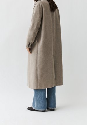 Osoba ubrana w długi beżowy płaszcz, niebieskie rozszerzane dżinsy i czarne buty, stojąca tyłem na jednolitym jasnym tle.