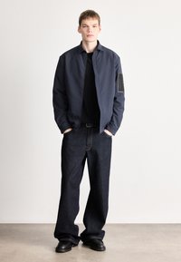 Lahka mornarsko modra jakna sproščenega kroja, kombinirana s temnimi kavbojkami. Ima črno teksturirano zasko na levi roki.