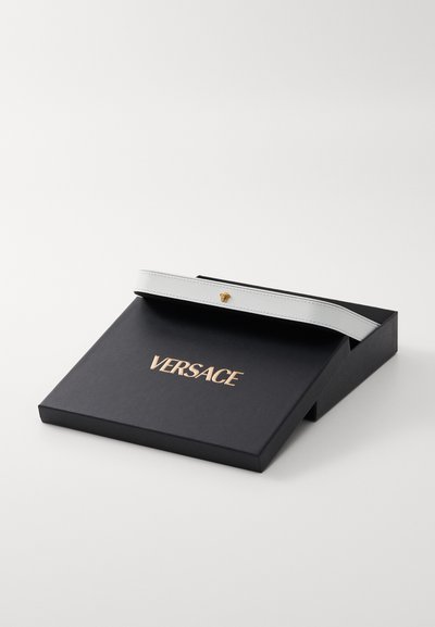 Baltas odinis diržas su auksiniu liūto aksesuaru juodame dėžėje, kurios viršus iš dekoruotas auksiniu „Versace“ logotipu, šiek tiek atidarytas.