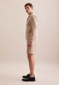 Giacca beige su misura con due tasche frontali, abbinata a pantaloni corti coordinati. Scarpe nere stringate. Texture liscia, palette di colori neutri.