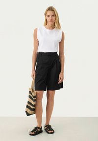 Vit ärmlös topp, svarta skräddarsydda shorts och svarta flätade sandaler. Håller en svart och beige randig vävd väska. Enkelt, modernt design.