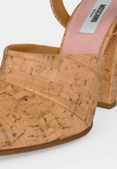Μπεζ σανδάλι με υφή φελλού, ανοιχτά δάχτυλα, φαρδιά λωρίδα και τακούνι μπλοκ, με ροζ εσωτερική σόλα με την επιγραφή Moschino Milano.