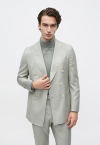 Mand iført lysegrå dobbeltradet jakkesæt med nålestriber og grå højhalset trøje, stående foran ensfarvet hvid baggrund.