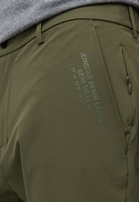 Pantalons vert olive au design épuré, avec une poche latérale, un marquage subtil et des coordonnées imprimées sur le tissu. Texture lisse.
