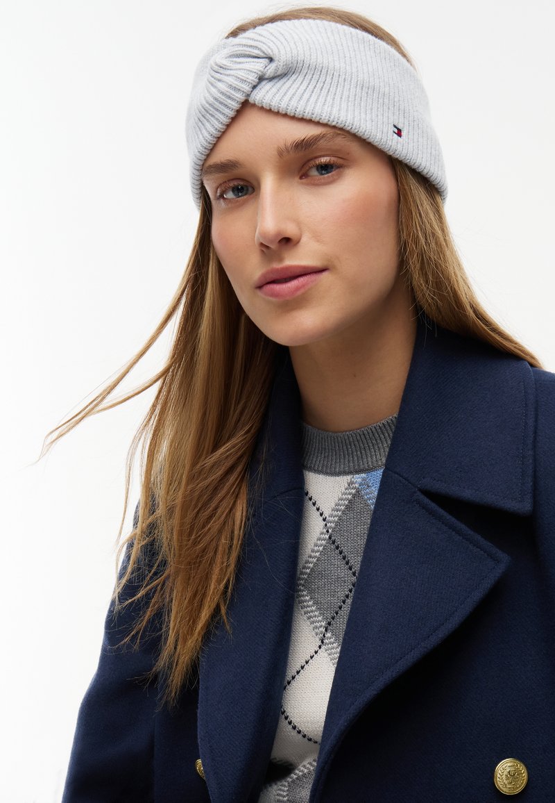 Jeune femme aux longs cheveux châtain clair portant un bandeau tricoté gris clair, un manteau bleu marine et un pull gris à motif losange, regardant légèrement sur le côté.