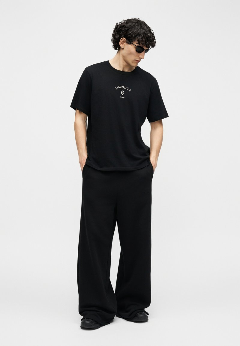 Junger Mann mit schwarzen Sonnenbrillen, der schwarze, übergroße Hosen und ein schwarzes T-Shirt mit weißem Text trägt und mit den Händen in den Taschen steht.