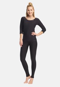 Smilende kvinde stående barfodet i sort tætsiddende langærmet top og leggings, med hånden på hoften, foran en ensartet lys baggrund.