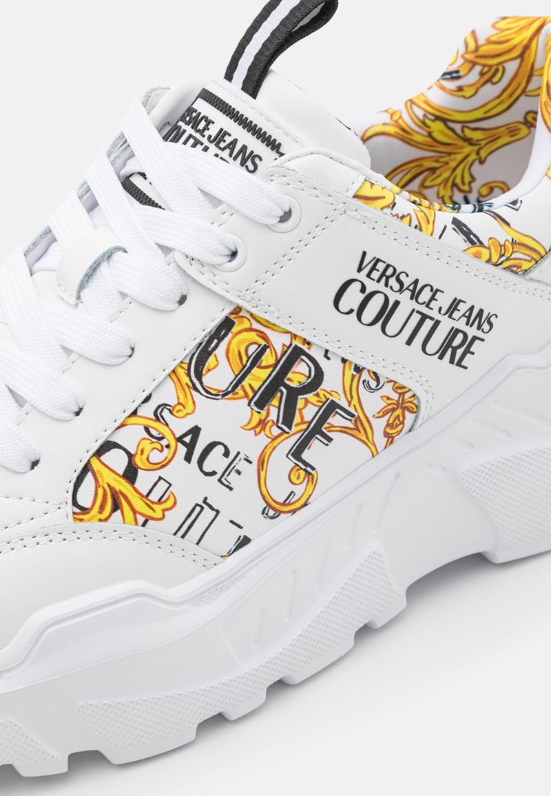 Sneaker bianco con motivi grafici in giallo e nero, dotata di una suola in gomma testurizzata, design con lacci e logo sul lato.