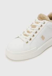 Hvite sneakers laget av teksturert lær, med beige faux fur-fôr, gulløyer, hvite snøringer og en liten preget logo.
