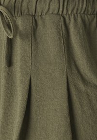 Detailní záběr na látku olivové zelené barvy s texturou, která má nabíraný pas a zavazovací tkaničku na levé straně.