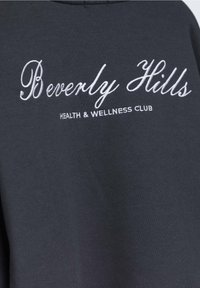 Sudadera negra con texto bordado en blanco que dice "Beverly Hills SALUD Y BIENESTAR CLUB." Diseño simple, textura suave.