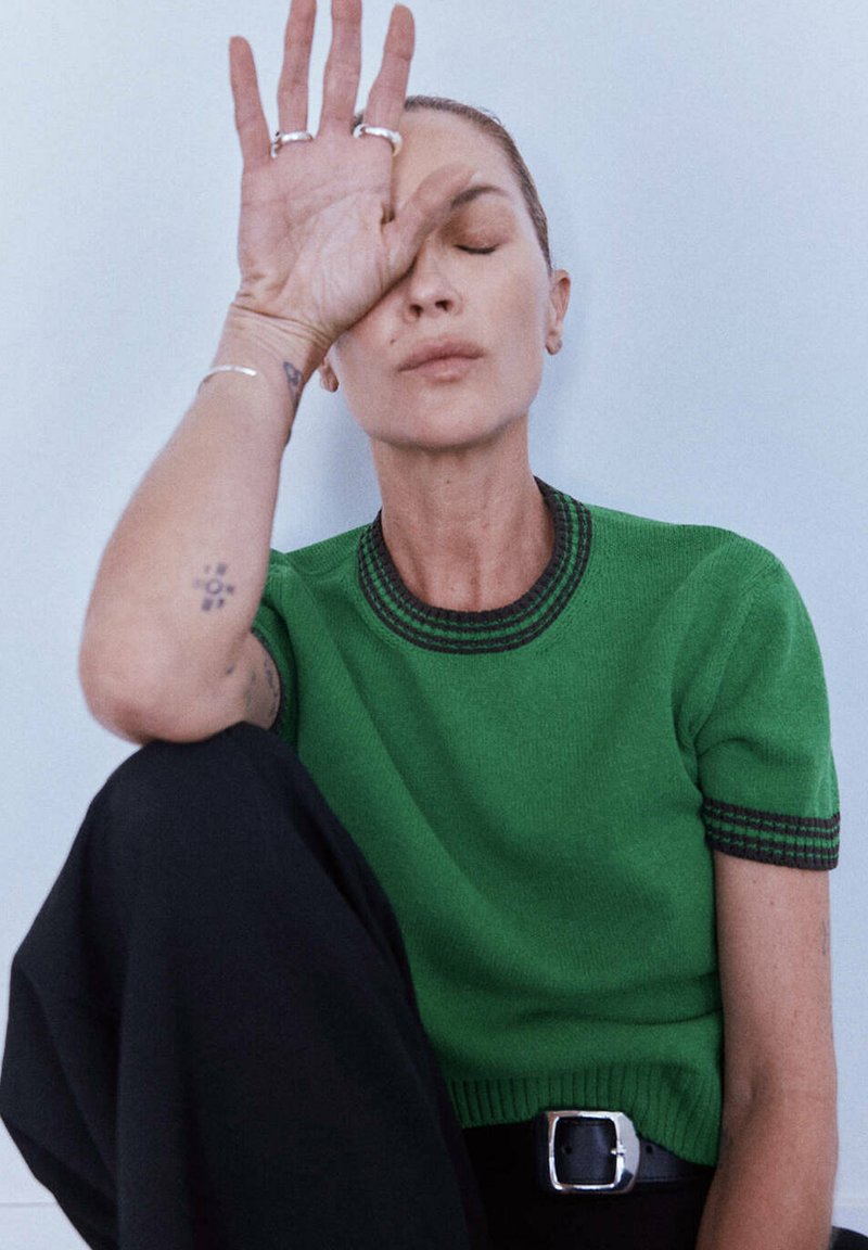 Femme portant un haut vert à manches courtes et un pantalon noir, assise les yeux fermés, levant la main vers le front, montrant des bagues et un tatouage sur son bras.