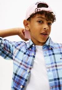 Overhemd met knopen in een blauw, roze en zwart plaid patroon, met eronder een wit T-shirt. Roze pet met "HILFIGER" geborduurd op de voorkant.