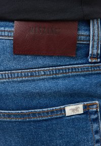 Niebieskie dżinsy z granatową skórzaną naszywką z napisem "MUSTANG" oraz małą białą metką z szarym logo zwierzęcia z tyłu.