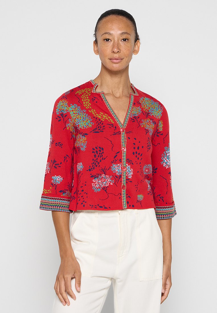 Kobieta w czerwonym swetrze zapinanym na guziki z niebieskimi i zielonymi wzorami kwiatowymi oraz białych spodniach z wysokim stanem, stojąca na jednolitym tle.