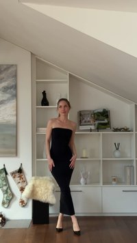 Zwarte strapless jurk met een aansluitende pasvorm en een zoom op knielengte, gedragen met zwarte hakken. Licht houten vloer en moderne witte planken op de achtergrond.
