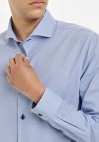 Vaaleansininen nappipaita, jossa on pieni, rakenteellinen kuvio. Paitassa on terävä kaulus, tummat napit ja säädettävät hihansuut.